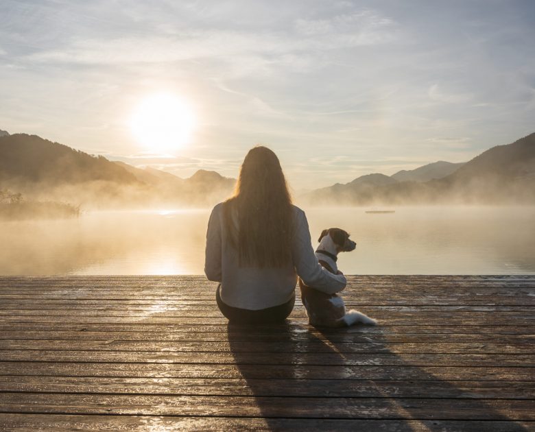 Rückansicht von einer jungen Frau und einem kleinen Terrier Hund auf einem Holzsteg an einem See. Sonnenaufgang, Nebel.