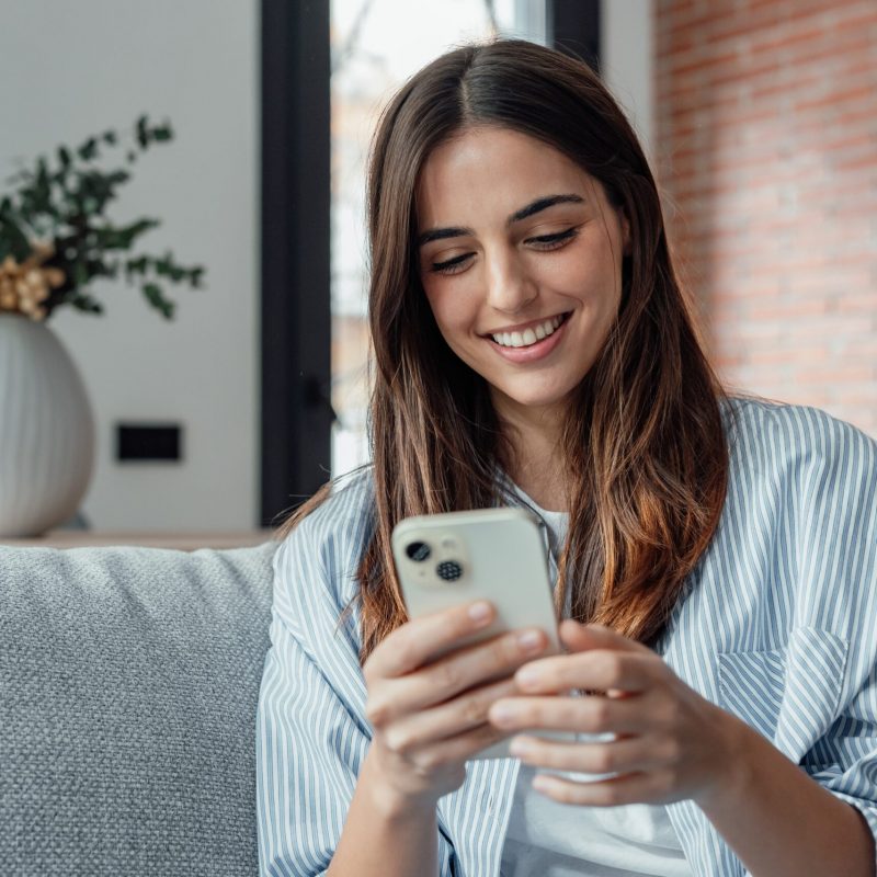 Smiling,Woman,Looking,At,Smartphone,Screen,,Sitting,On,Couch,,Enjoying